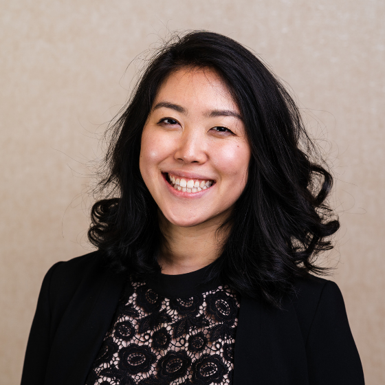 Smiling woman with wavy, mid-length black hair, wearing a black lace top with a black blazer, against a solid beige background.