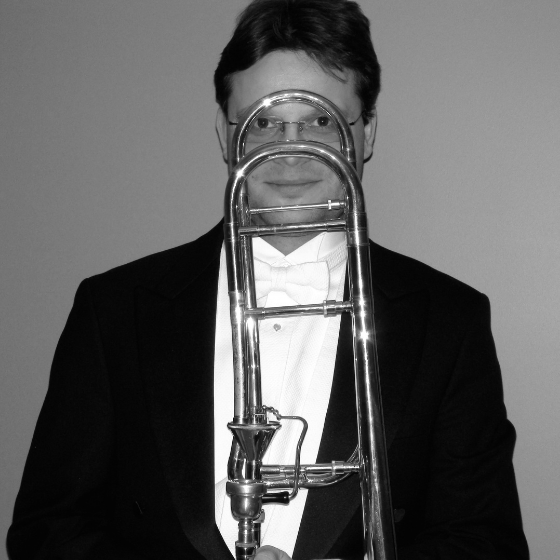 Monochrome image of a man in a suit, smiling and holding a trombone in front of his face.