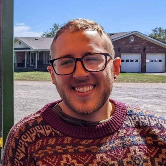 Smiling man with glasses, wearing a knit sweater, pictured in front of the backyard of a house.