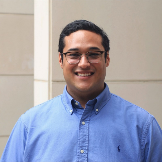 Smiling man with short dark brown hair, wearing glasses and a blue long sleeved top with a collar, against a white wall.