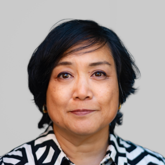 Woman with short black hair, wearing a black and white striped blouse, pictured against a solid grey background.