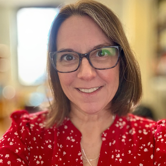 Smiling woman with shoulder length brown hair, wearing glasses and a red top, against a blurred room background.
