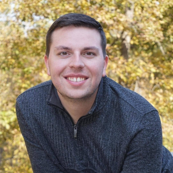 A smiling man with short brown hair, wearing a dark grey sweater, leaning forward slightly, with a backdrop of autumn trees.