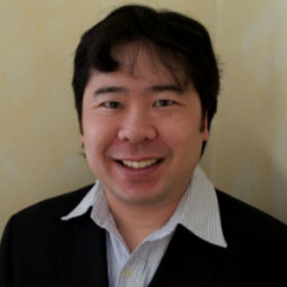 Smiling man with short black hair, wearing a black blazer and a white striped shirt, against a neutral wall background.