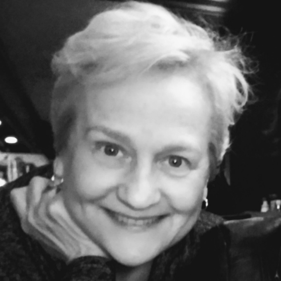 Monochrome picture of a smiling woman with short white hair, against a dark background.