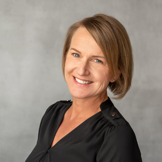 Smiling woman with short dark blonde hair, wearing a black top, against a grey background.