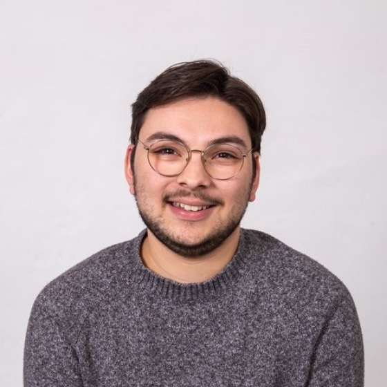 Smiling man with short brown hair, wearing glasses and a dark grey sweater, against a solid off-white background.