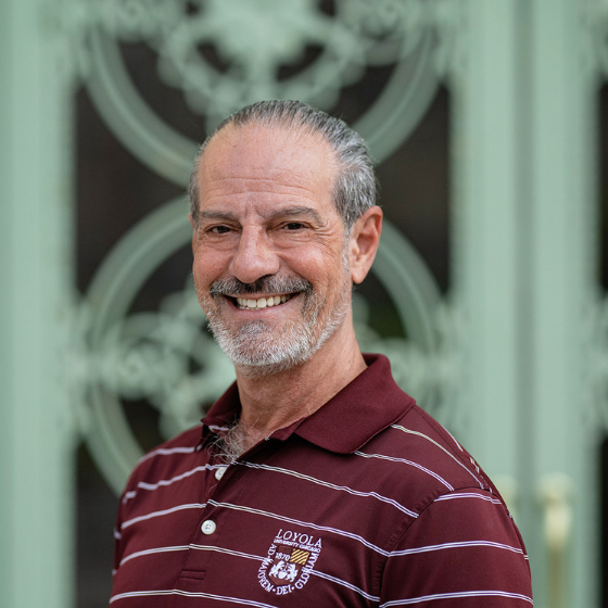Smiling man with grey hair pulled back into a ponytail, wearing a striped maroon shirt with the Loyola logo on it, pictured outside in front of an ornate gate.