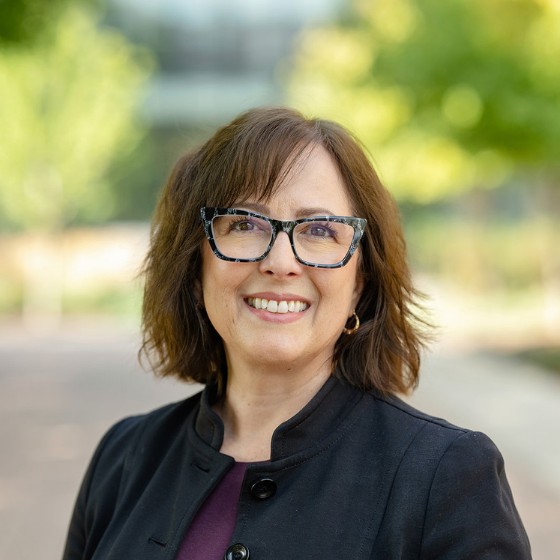 Square headshot of a brunette white woman with glasses, with a college campus in the background