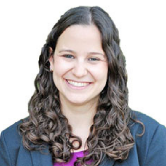 A smiling woman with medium length, wavy brown hair, wearing a dark blue blazer over a magenta top, behind a solid white background.