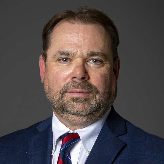 Man with short brown hair smiling faintly and wearing a dark blue suit jacket, white top, and a red tie, in front of a solid dark grey background.