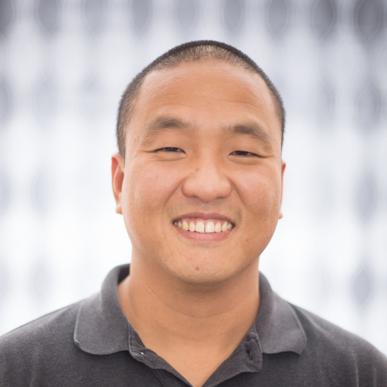 Smiling man with a shaved head, wearing a dark grey shirt with a collar, in front of a checkered curtain.