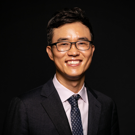 Smiling man with short dark brown hair, wearing glasses, a black suit jacket, and a white top with a dark grey tie, against a solid black background.