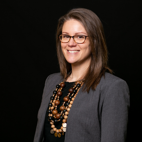 Woman with medium brown hair, smiling and wearing glasses, a dark grey suit jacket, a black top, and several chunky necklaces, against a solid black background.