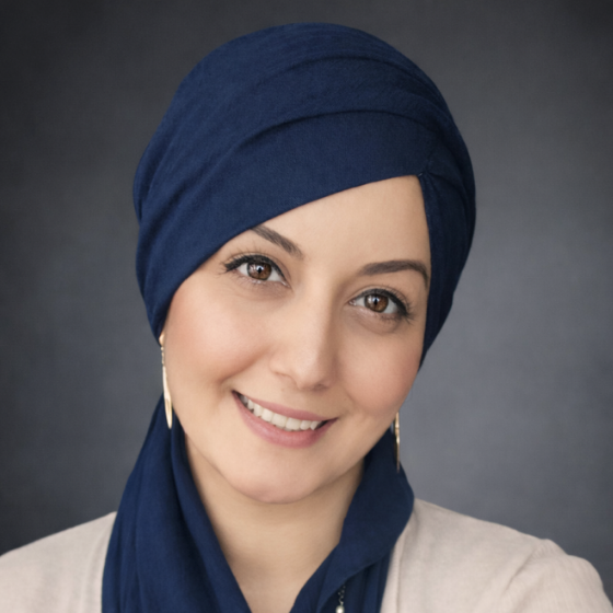 Smiling woman with a navy blue headscarf, wearing a white top, against a dark grey background.