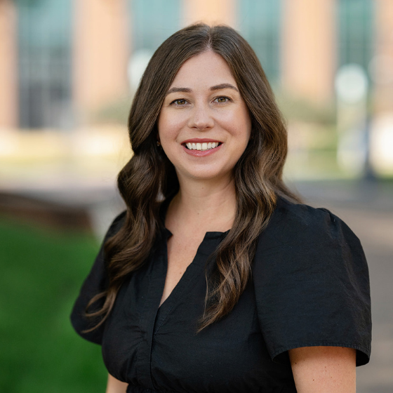 Woman smiling with long, wavy brown hair, wearing a black blouse, standing in front of a Loyola University Chicago campus building.