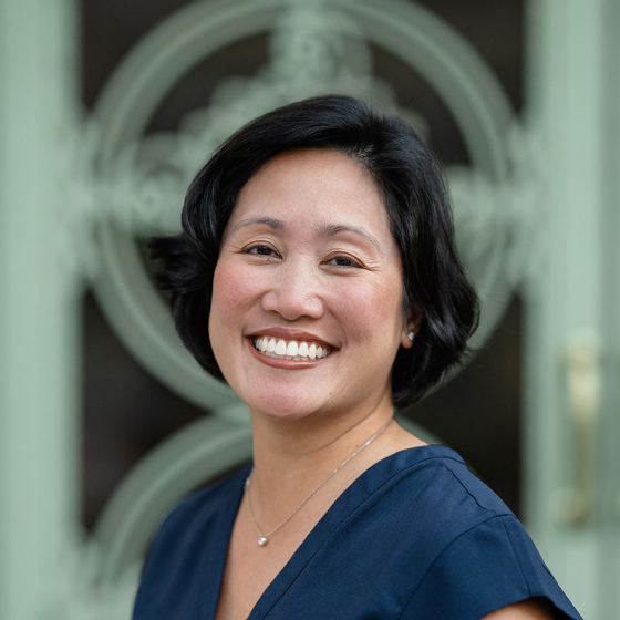 Smiling woman with her black hair in a short bob, wearing a navy blue top, standing in front of a building gate.