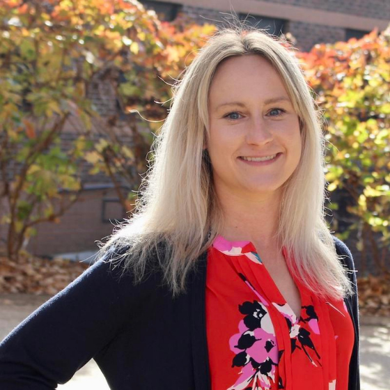 Smiling woman with shoulder length blonde hair, wearing a dark grey cardigan over a floral red top, with greenery in the background.