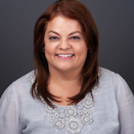 A woman smiling with long brown hair, wearing a light grey blouse, against a solid dark grey background.