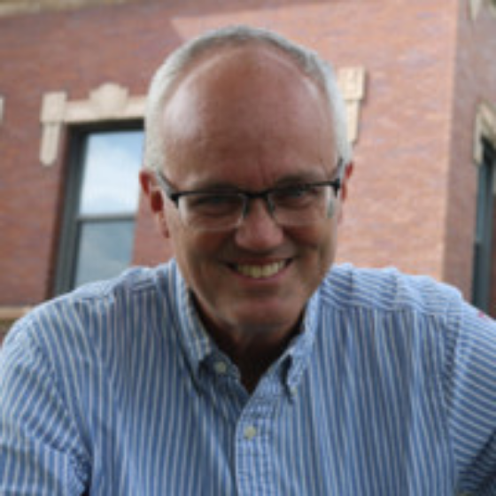 Bald man smiling, wearing glasses, and a light blue button-up top, with a building in the background.