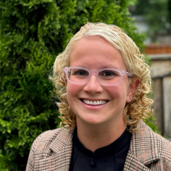 Woman smiling with curly blonde hair, wearing glasses, a gingham tan blazer over a black top, with greenery in the background.