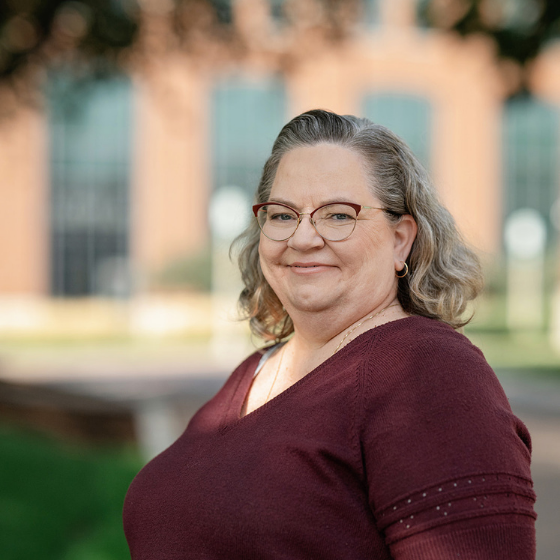Smiling woman wearing glasses and a maroon colored top, pictured outside in front of a campus building.