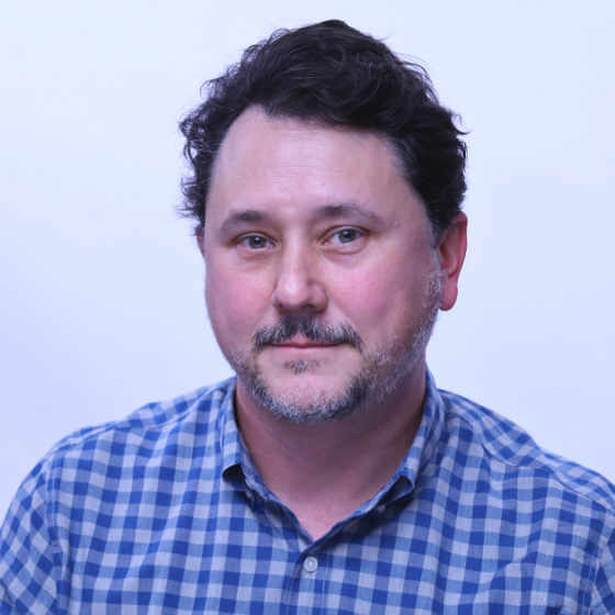 A man with short, dark brown hair, smiling slightly, wearing a checkered blue button-up top, against a solid light grey background.