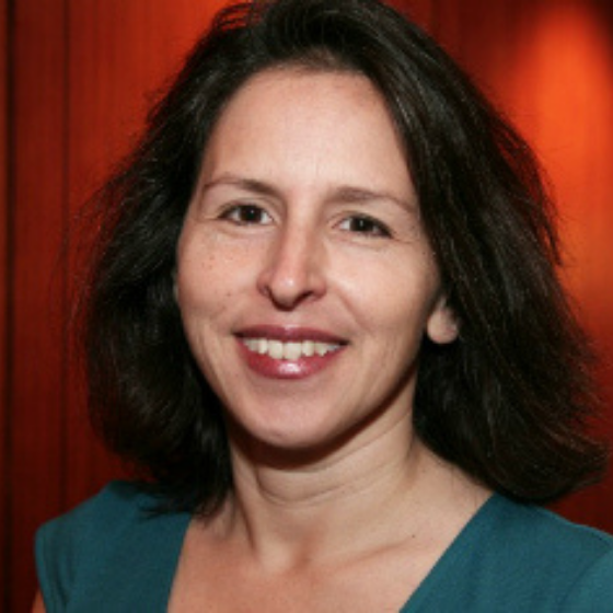 A woman smiling with shoulder-length brown hair, wearing a teal top, against a dark red wall.