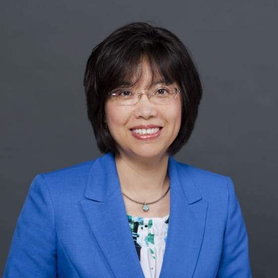 A woman smiling with short black hair, wearing glasses, a necklace, and a blue blazer over a floral light blue top, against a solid dark grey background.