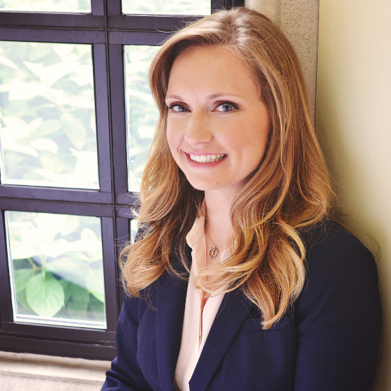 A woman smiling with long, wavy blonde hair, wearing a dark blue blazer over a light pink top, leaning against a windowsill.