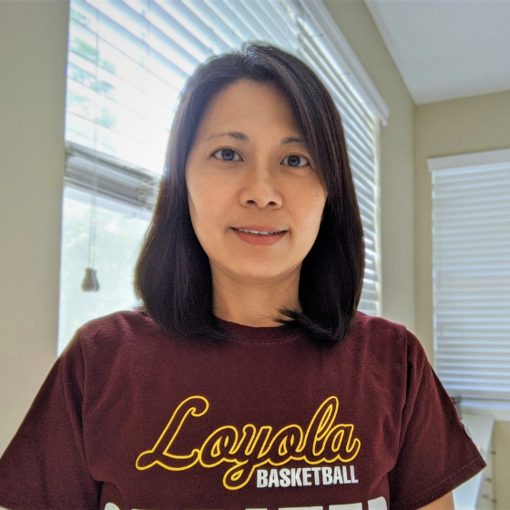 A woman with shoulder-length brown hair, smiling slightly, wearing a maroon Loyola University Chicago top, with a bright sunroom in the background.