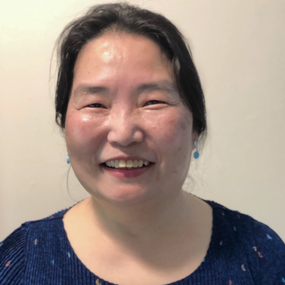 A woman smiling with short black hair, wearing a dark blue top, against an off-white background.