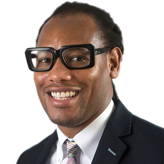 Smiling man with black hair pulled back into locs, wearing thick-rimmed black glasses, a suit jacket, and plaid patterned tie, against a solid white background.