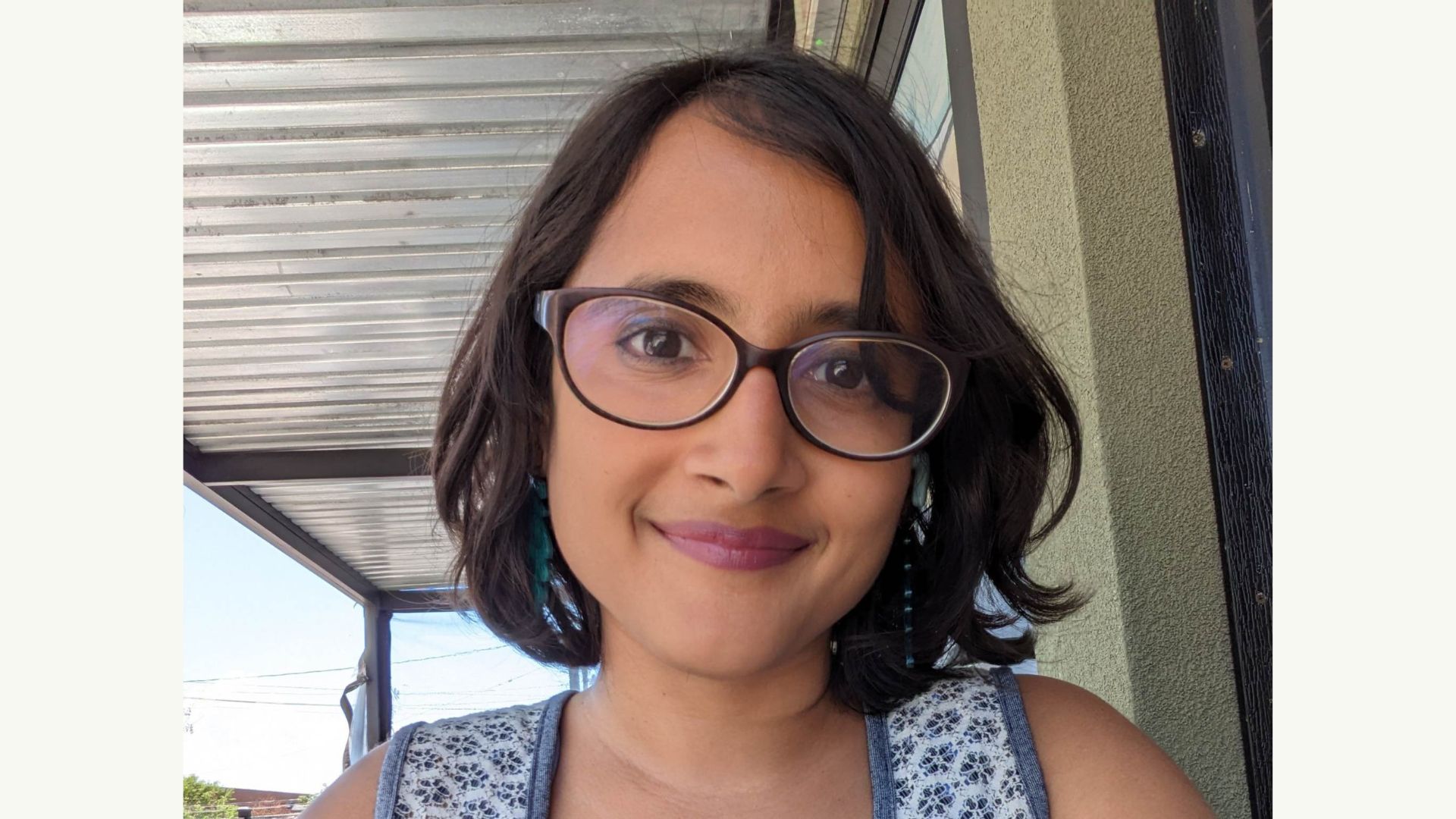 Woman with short dark hair and glasses smiling at the camera outdoors under a covered walkway.