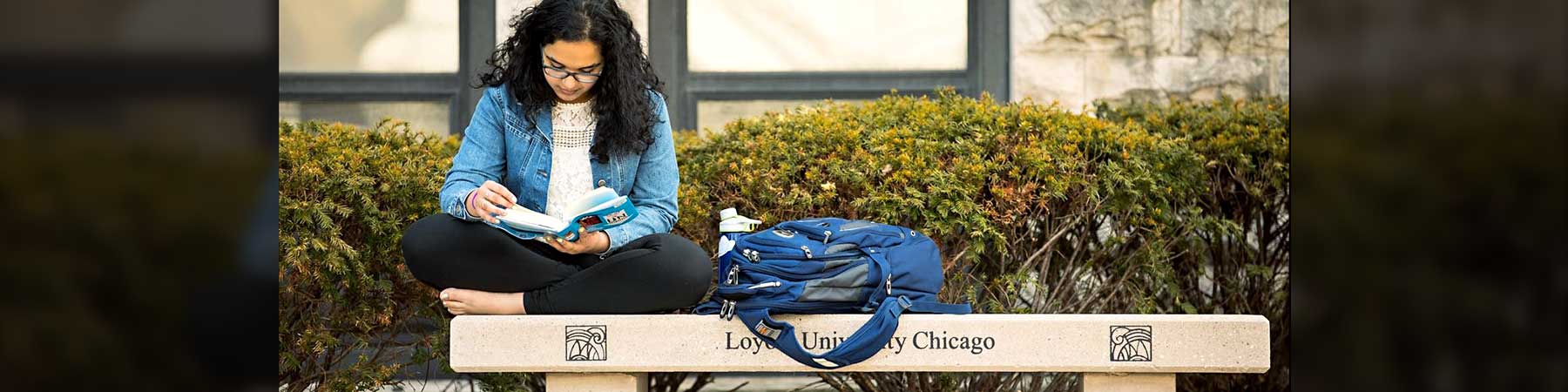 Student sitting on a campus bench reading a book, with a backpack beside them.