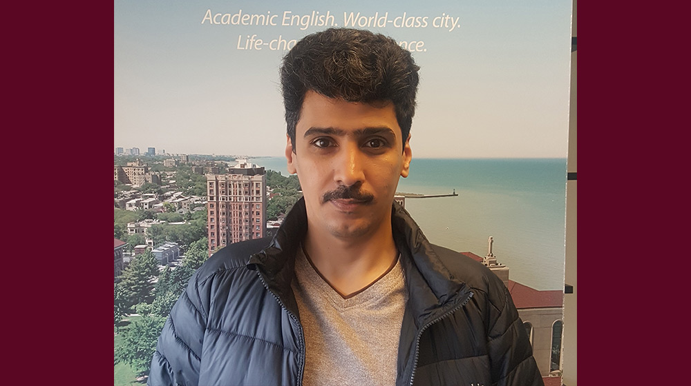 Ahmed wearing a winter jacket standing in front of a ESL Program banner