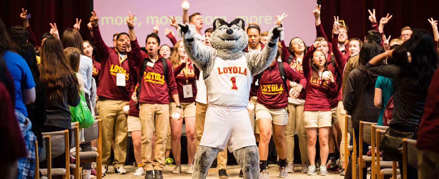 Loyola mascot leads cheering students at campus event in auditorium.