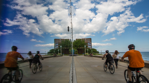 Students ride past the Information Commons building where the blue sky, clouds, and lake are mirrored in its windows.