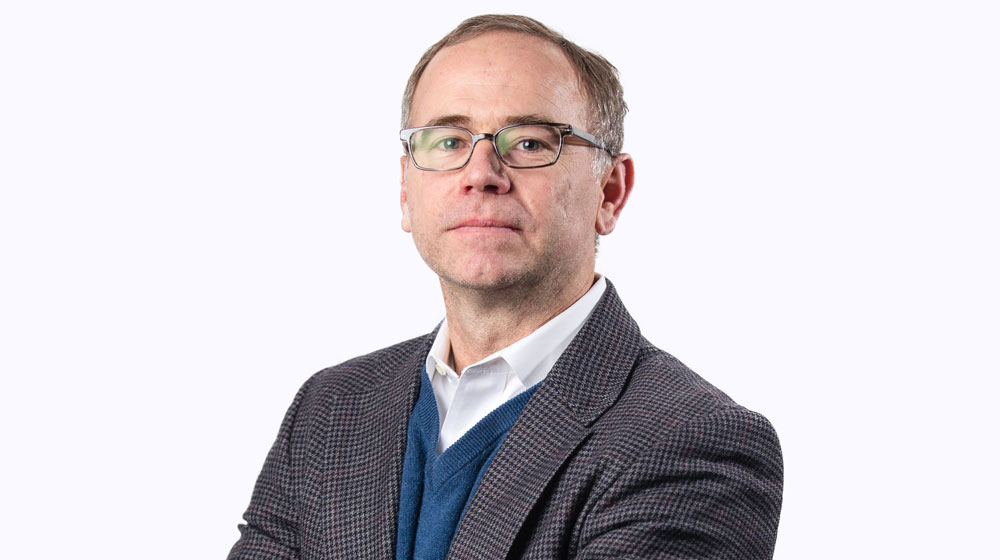 Headshot of a man with glasses wearing a patterned blazer and layered sweater over a collared shirt against a light background