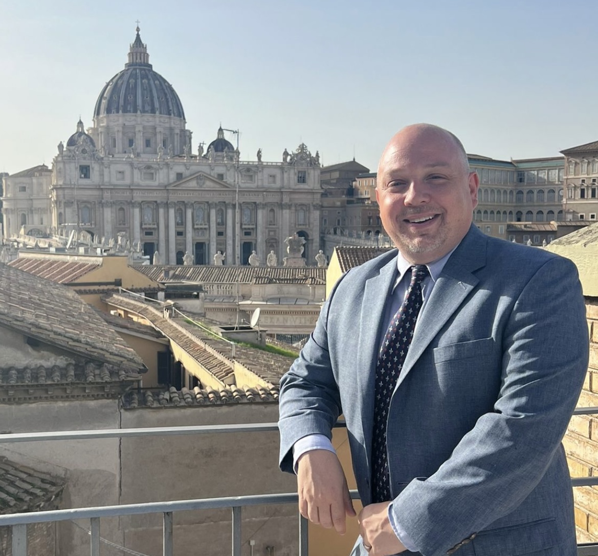 Man in a suit smiling on a terrace with St. Peter’s Basilica and Vatican City buildings behind him