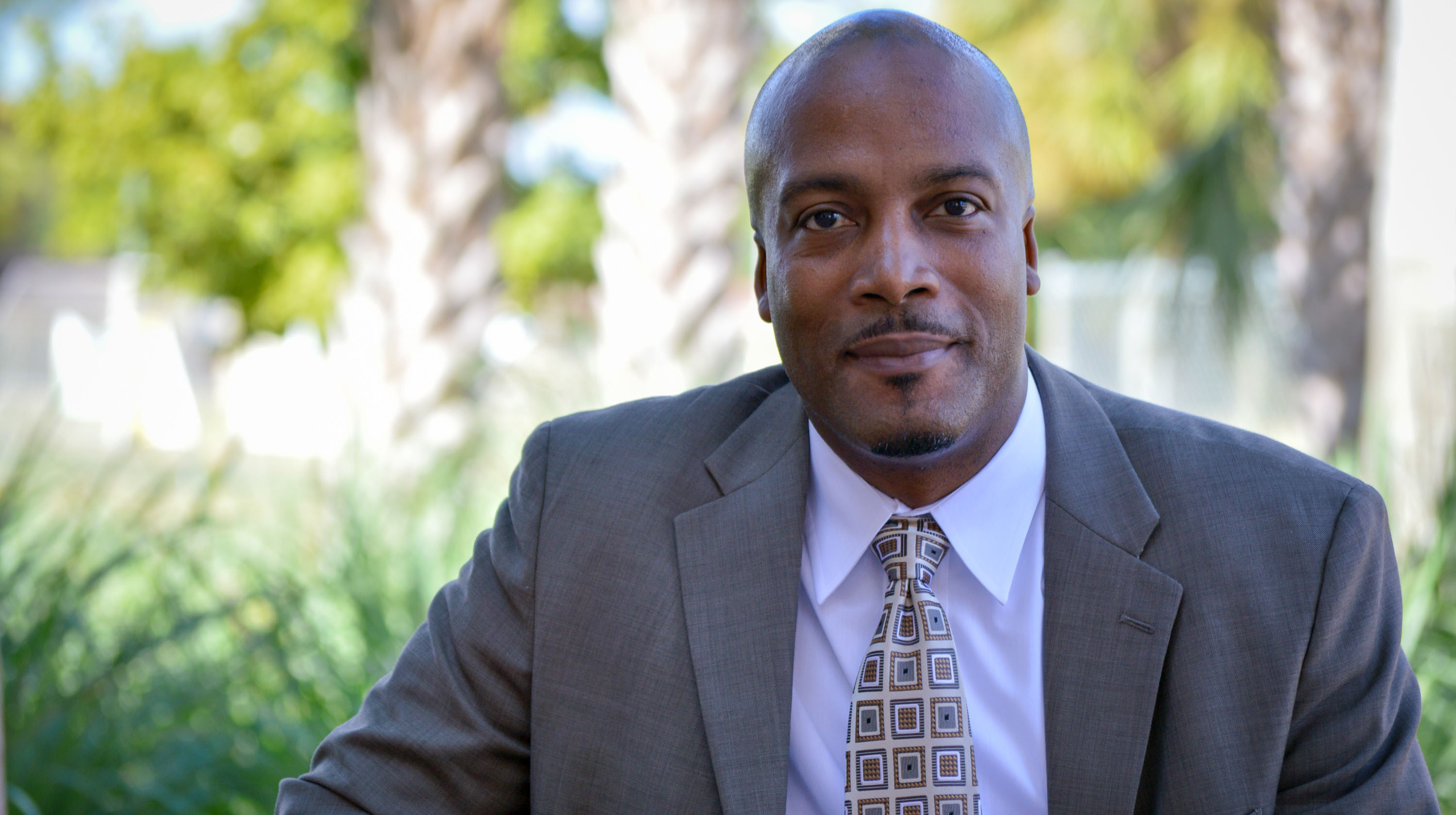 Man in a suit and patterned tie sitting outdoors, with blurred greenery and palm trees behind him