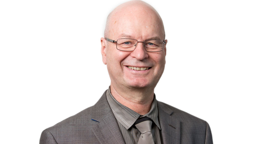 Headshot of a smiling man with glasses, wearing a gray suit and tie against a light background