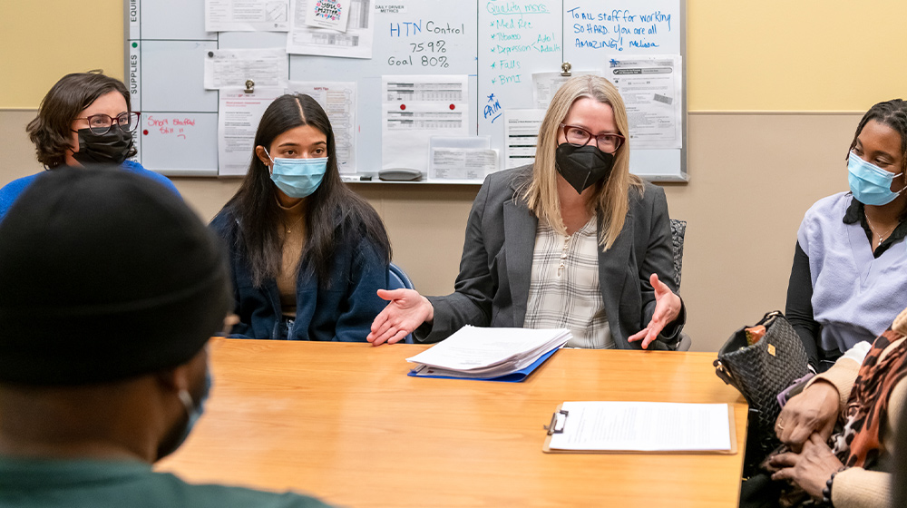photo of professor and students sitting around a table. They appear to be talking but all have masks on.