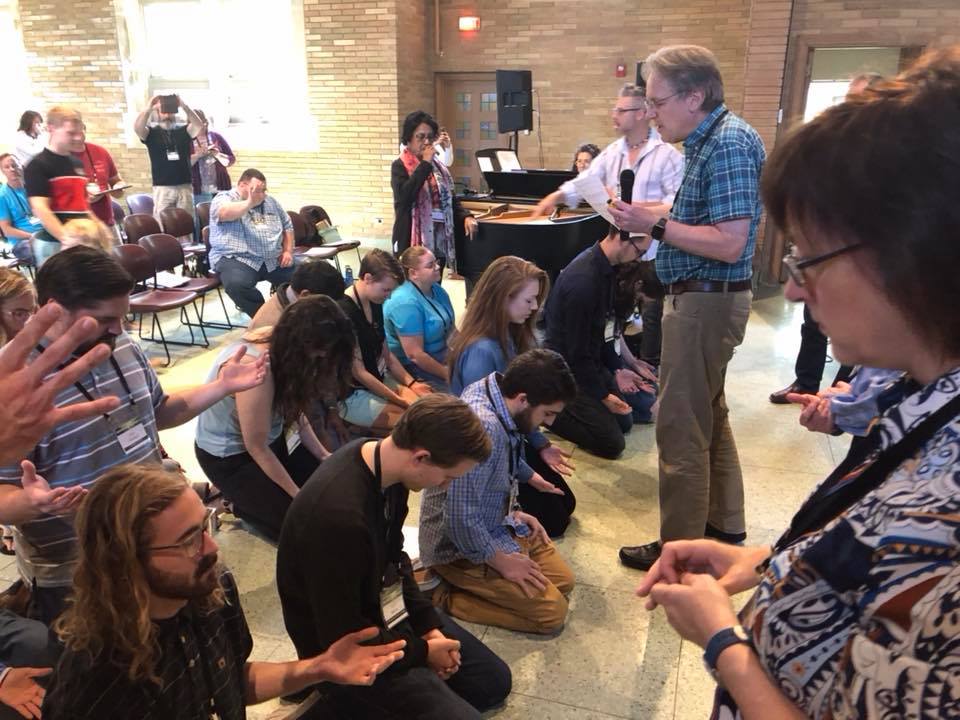 A worship group gathers in a room with many of them on their knees and praying