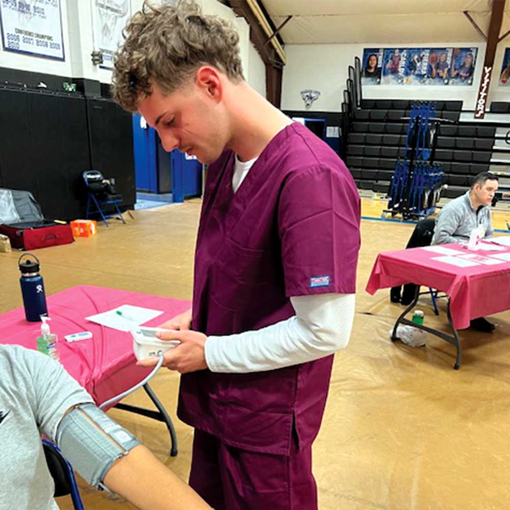 Person in maroon scrubs checks blood pressure of another individual seated at a table during a health screening event.