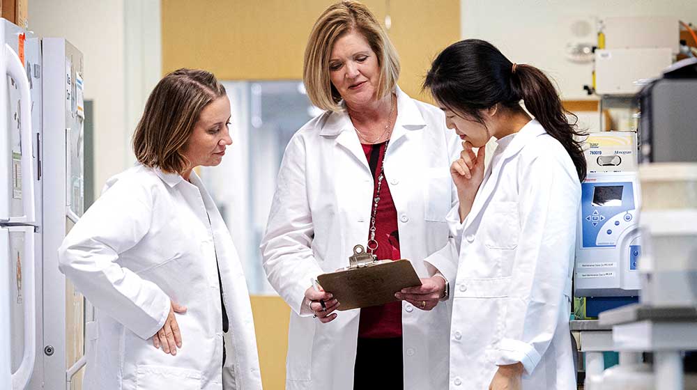 Three individuals in white lab coats stand in a laboratory, reviewing notes
