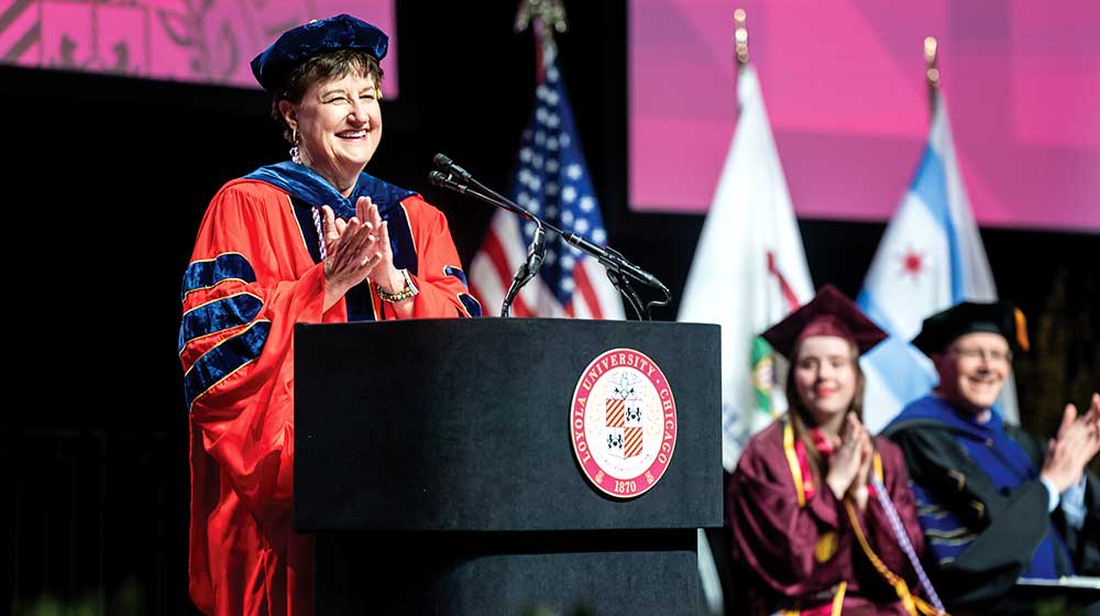 Dean Lorna Finnegan stands at a podium in academic regalia, clapping during a commencement ceremony with flags in background.
