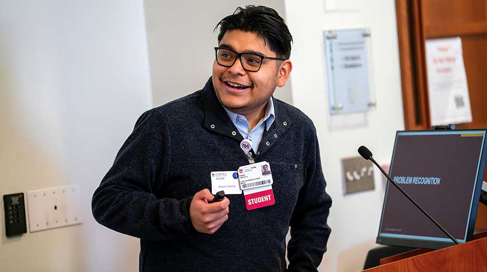 Student wearing a dark sweater and ID badge presents at a podium with a microphone and screen displaying “Problem Recognition.”