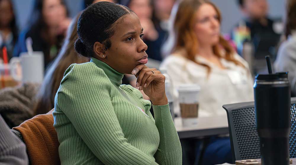 Person wearing a green ribbed sweater sits attentively at a conference table with a coffee cup and tumbler in view.