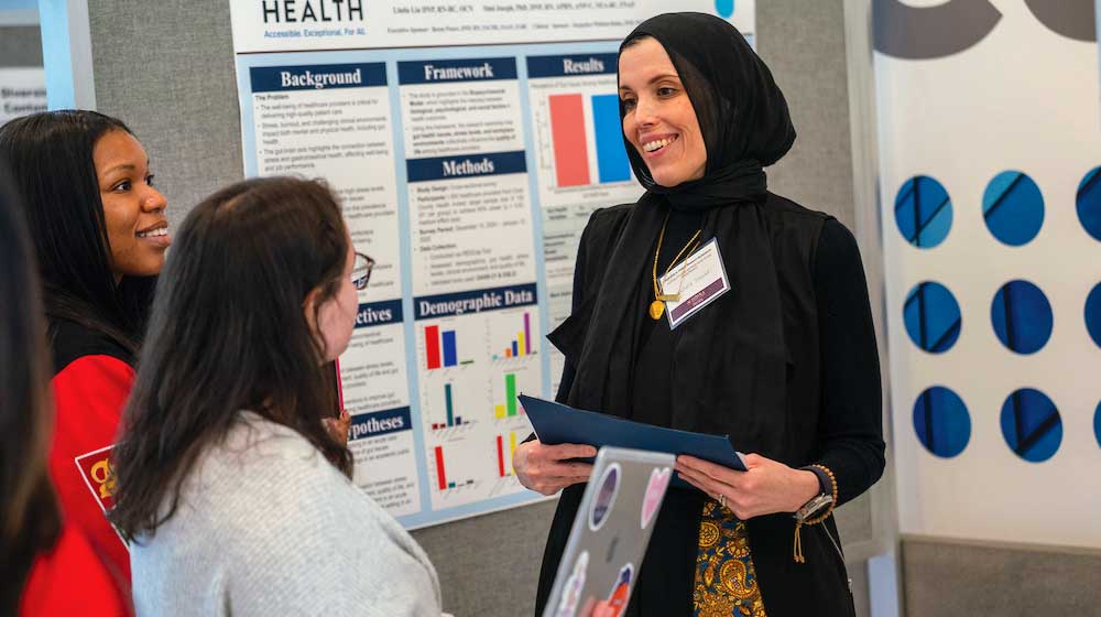 A person wearing a black headscarf and black outfit with a patterned gold design holds a folder while presenting research at a poster session.